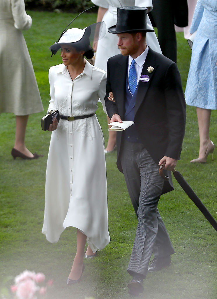 Meghan Markle îmbrăcată cu o rochie albă și o pălărie albă pe cap la brațul Prințului Harry îmbrăcat cu frac și joben