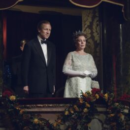 Olivia Colman îmbrăcată cu o rochie albă elegantă și o coroană pe cap în rolul Reginei Elisabeta a II-a