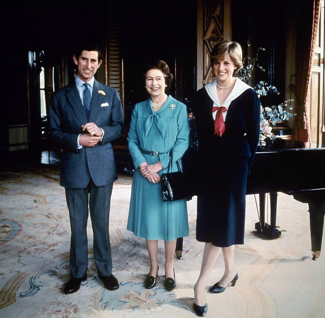 Prințesa Diana îmbrăcată într-o rochie neagră pozează într-o sufragerie alături de Regina Elisabeta îmbrăcată cu un costum albastru și de Prințul Charles îmbrăcat cu un costum gri