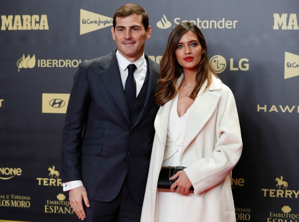 Sara Carbonero îmbrăcată ăntr-o rochie albă și palton alb pe covorul roșu alături de Iker Casillas