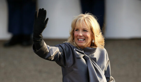 Jill Biden salută mulțimea îmbrăcată într-un palton gri cu fundă și mănuși negre