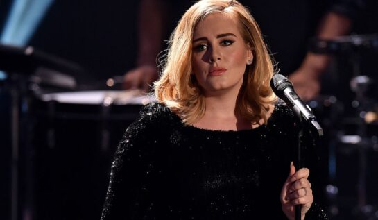 Adele pe scenă cu microfonul în mână îmbrăcată cu o eochie neagră sclipitoare