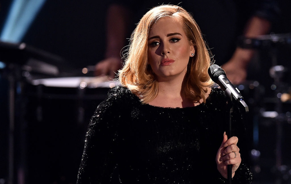 Adele pe scenă cu microfonul în mână îmbrăcată cu o eochie neagră sclipitoare
