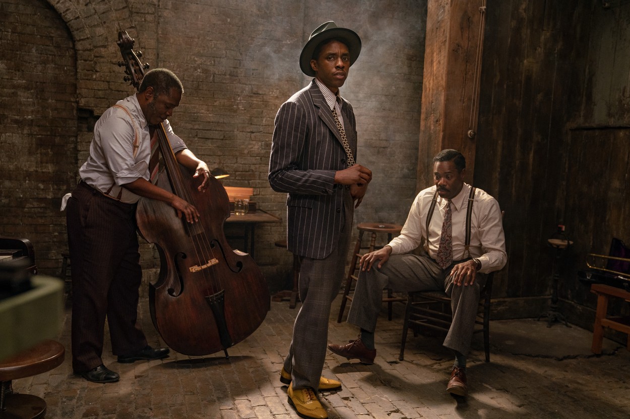 Chadwick Boseman în Ma Rainey’s Black Bottom îmbrăcat cu un costum gri și o pălărie alături de doi cântăreți de jazz într-un decor de epocă