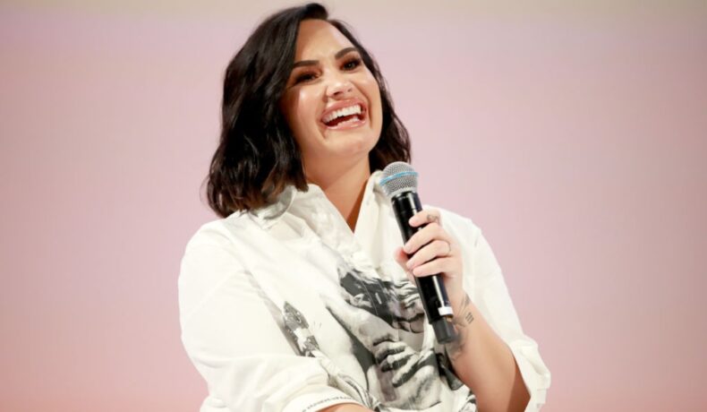 Demi Lovato zâmbește în timp ce vorbește la microfon îmbrăcată cu o cămașă albă cu aplicații negre
