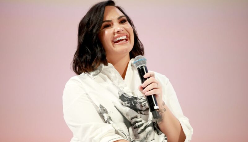 Demi Lovato zâmbește în timp ce vorbește la microfon îmbrăcată cu o cămașă albă cu aplicații negre