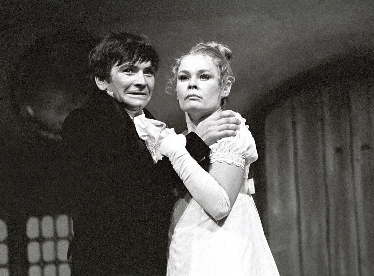 Fotografie alb-negru cu Judi Dench tânără cu părul prins în coc și o rochie albă care ține în brațe un actor pe o scenă de teatru