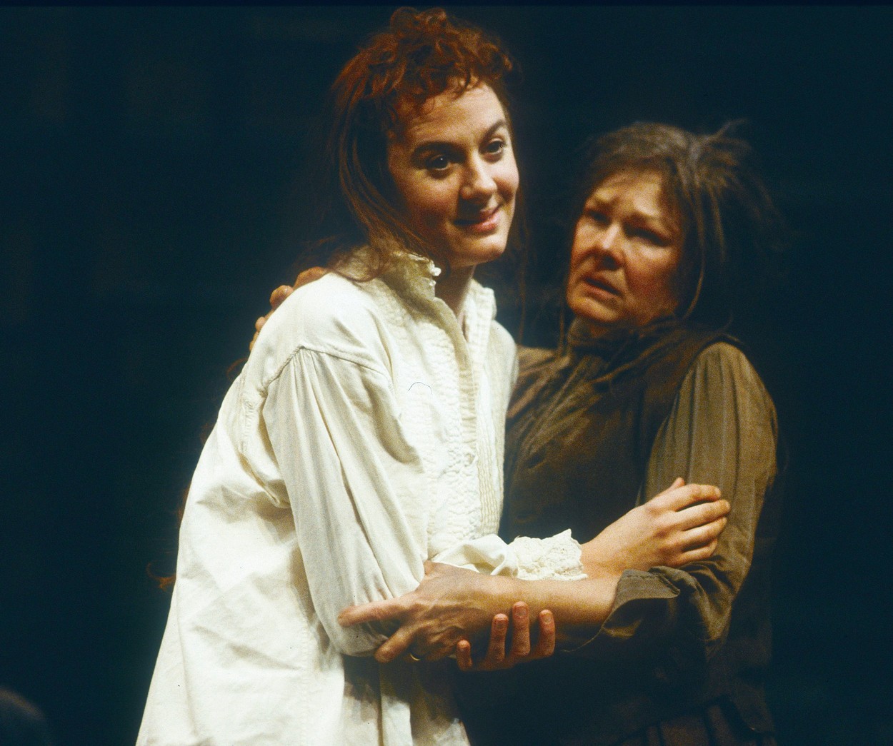 Judi Dench cu părul lung șaten și haine maro ține în brațe un actor pe o scenă de teatru