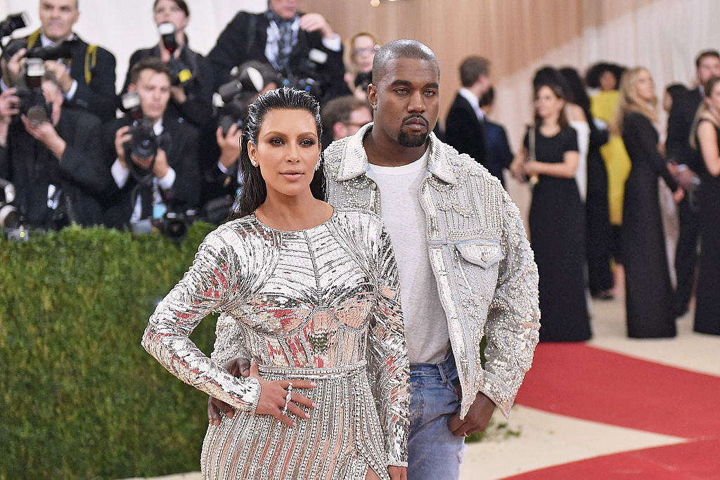 Kanye West îmbrăcat cu o jachetă argintie cu aplicații stă în spatele lui Kima Kardashian îmbrăcată cu o rochie argintie pe covorul roșu la Met Gala