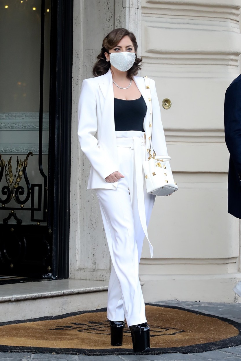 Lady Gaga îmbrăcată cu sacou și pantaloni albi și cizme negre cu toc foarte înalt în fața unui hotel
