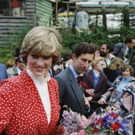 Lady Diana, alături de Prințul Charles, poartă o rochie roșie, cu buline și guler încrețit, și ține în mână un buchet de flori.