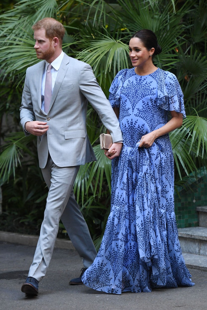 Prințul Harry îmbrăcat cu un costum gri deschis o ține de mână pe Meghan Markle îmbrăcată cu o rochie albastră lungă