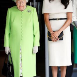 Regina Elisabeta a II-a, îmbrăcată într-un costum verde fistic, alături de Meghan Markle, care poartă o rochie de culoare albă, accesorizată cu o curea subțire neagră.
