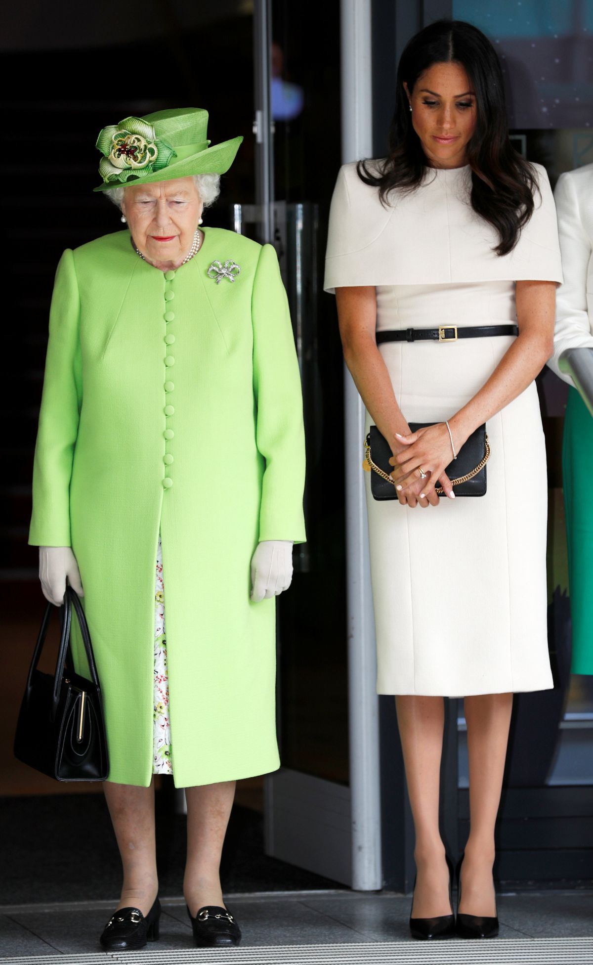 Regina Elisabeta a II-a, îmbrăcată într-un costum verde fistic, alături de Meghan Markle, care poartă o rochie de culoare albă, accesorizată cu o curea subțire neagră.