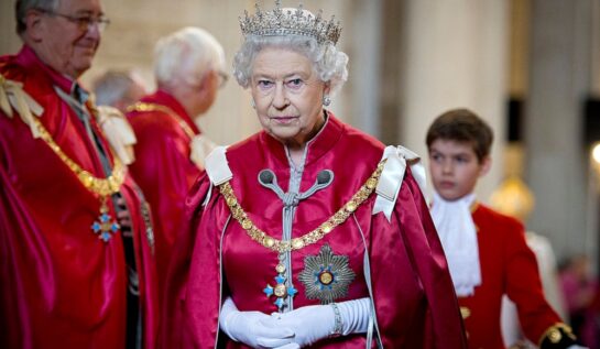 Regina îmbrăcată cu un costum roșu din satin cu capă și coroană pe cap la un eveniment