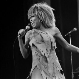 Tina Turner, fotografiată pe scenă în 1983, în timp ce ține un microfon în mână și poartă o rochie scurtă, sfâșiată.
