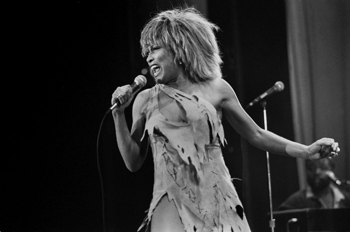 Tina Turner, fotografiată pe scenă în 1983, în timp ce ține un microfon în mână și poartă o rochie scurtă, sfâșiată.
