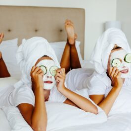 Două femei, la spa. Ambele sunt pe un pat cu lenjerie albă, cu prosoape albe pe cap, cu măști albe pe față și cu castraveți proaspeți pe ochi.