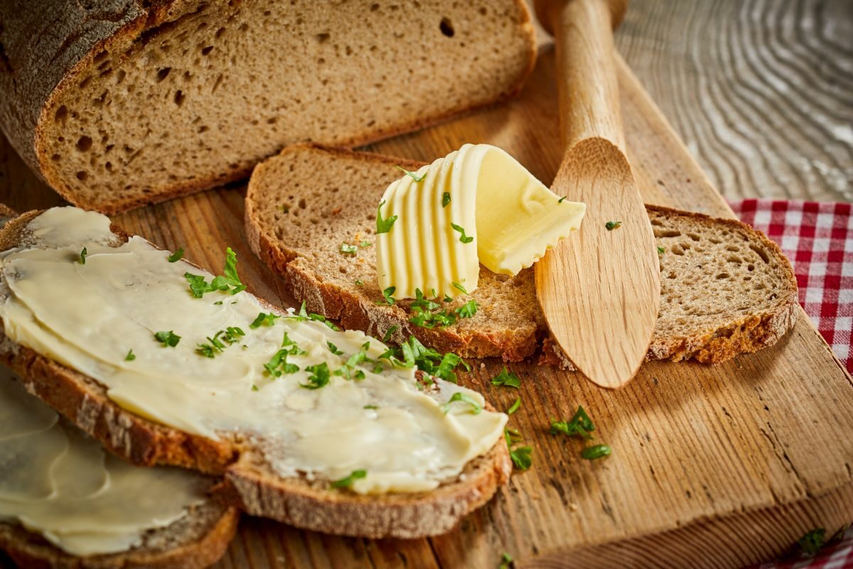 Felie de pâine, unsă cu unt, care are deasupra pătrunjel, lângă o pâine tăiată și un cuțit