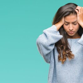 O tânără femeie, ce poartă un pulover albastru de iarnă, își ține tâmplele îmn palmă, din cauza unei dureri de cap