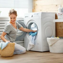 O femeie, în pantaloni albi și tricou gri, utilizează mașina de spălat