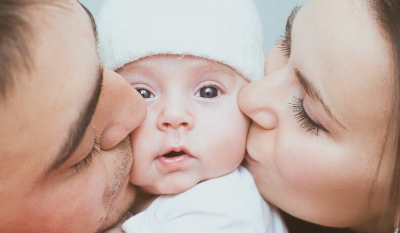 Mama și tata își sărută bebelușul, fiecare pe câte un obraz