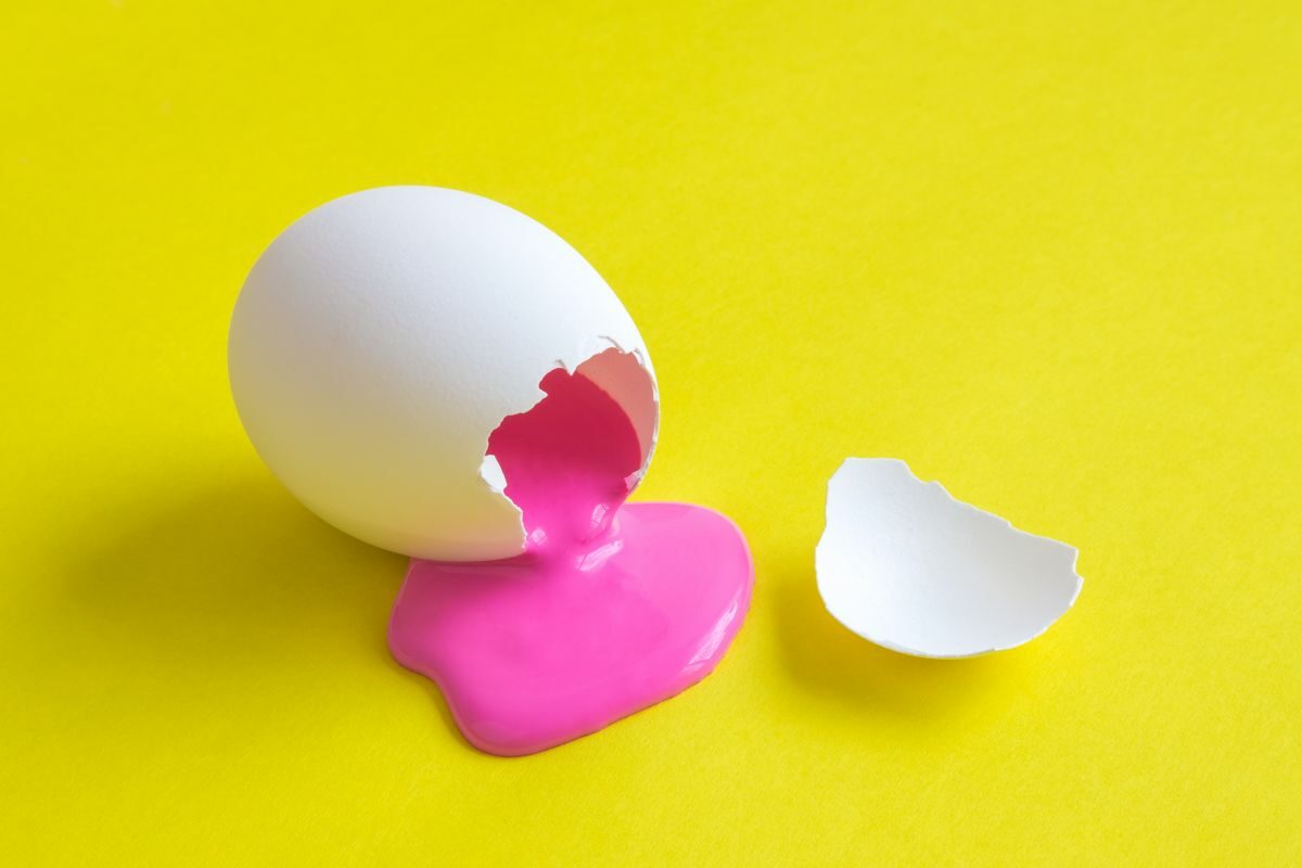 Un ou alb, spart, din care curge vopsea roz, pe un fundal galben
