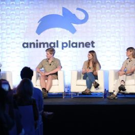 Irwin Bindi cu familia sunt într-un platou de emisiune. În stâng este prezentatorul. ÎN spate au logo-ul de la Animal Planet. toți poartă cămăși deschise.
