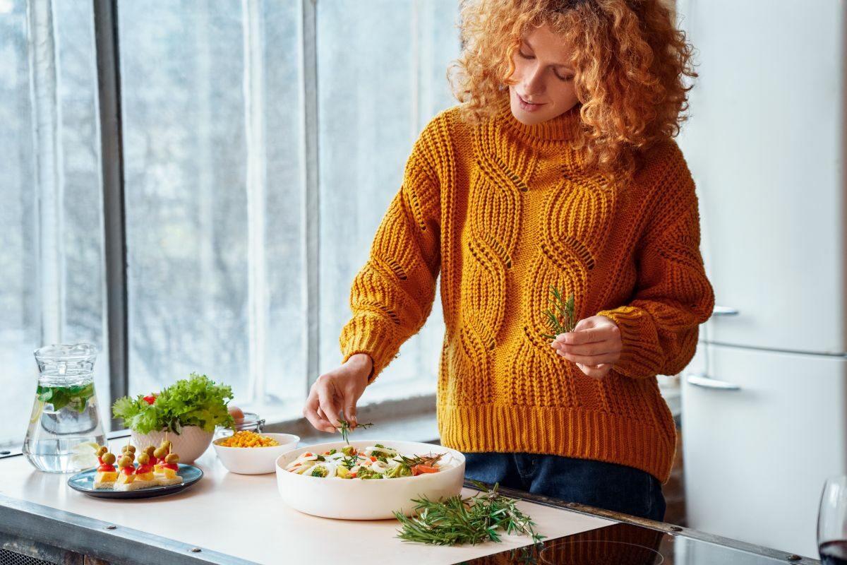 O femeie fericită,îmbrăcată într-un pulover galben, ornează o salată, într-o bucătărie