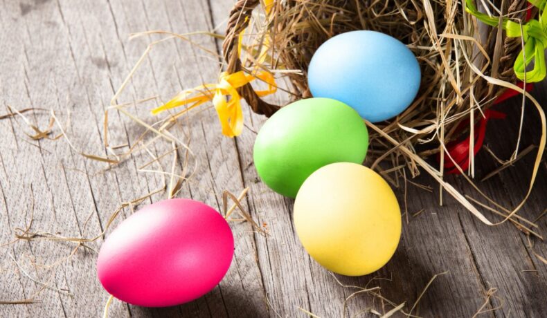 Patru ouă colorate, de Paște, lângă un coș maro, de răchită