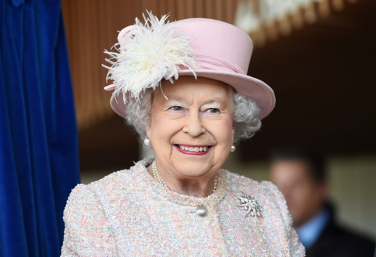 Regina Elisabeta cu o pălarie albă cu pene într-un costum alb în timp ce poartă una din broșele sale celebre numită steaua jardinei