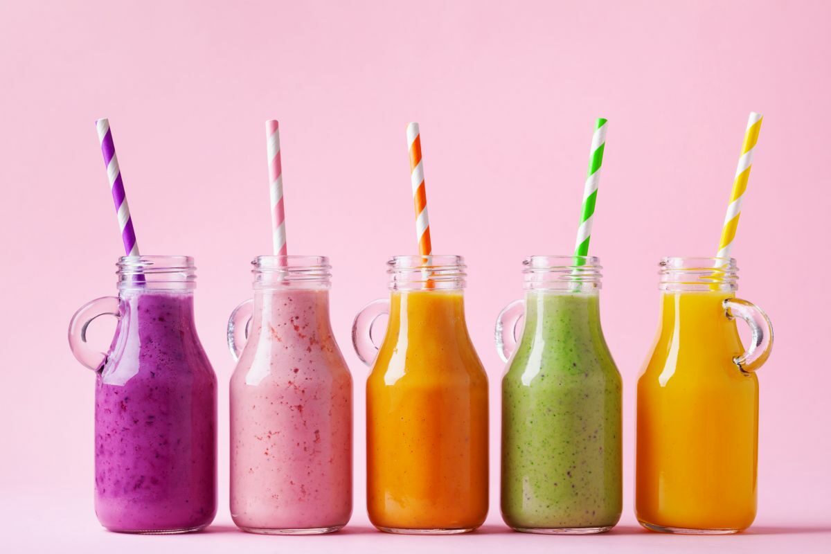 Cinci sticle ce conțin shake-uri colorate de fructe, pe un fundal roz
