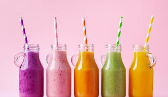 Cinci sticle ce conțin shake-uri colorate de fructe, pe un fundal roz