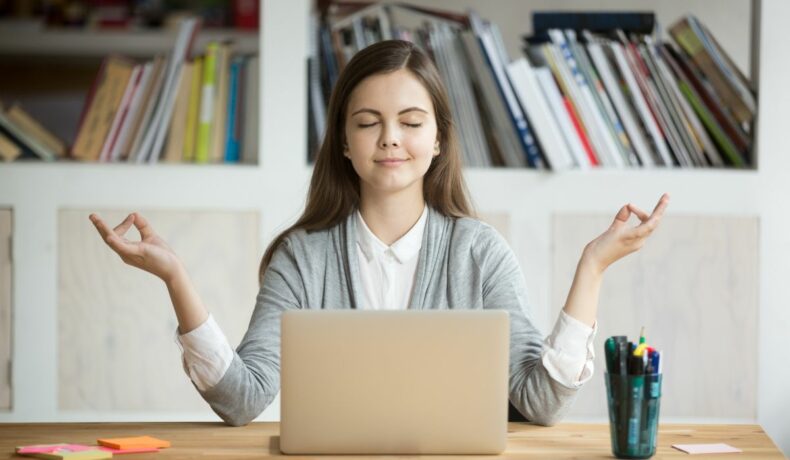 Femeie care meditează relaxată, cu ochii închii și surâzând, în fața laptopului