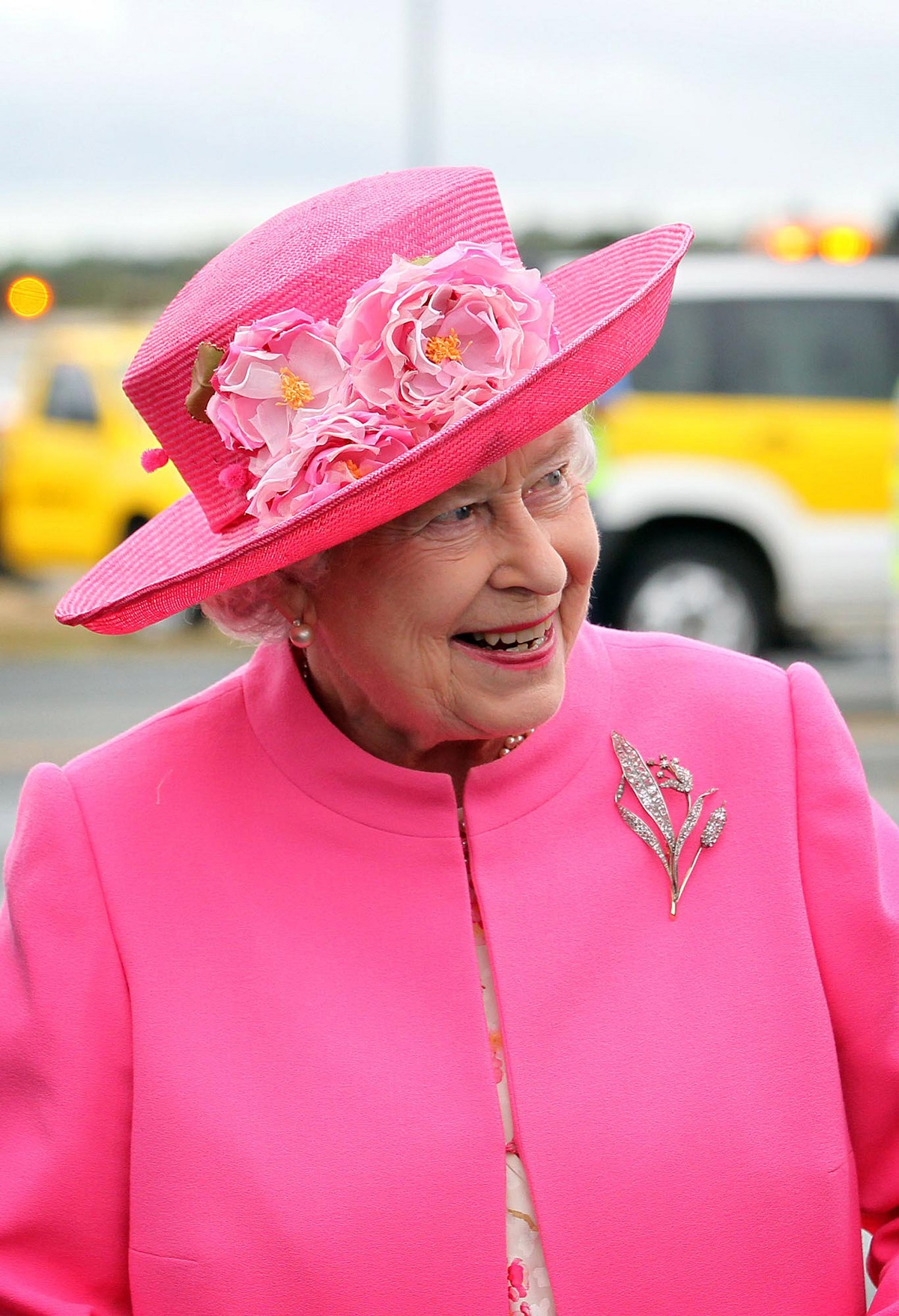 Regina Elisabeta cu o pălărie și un costum roz purtând în piept brosa sa Crinul Londrei