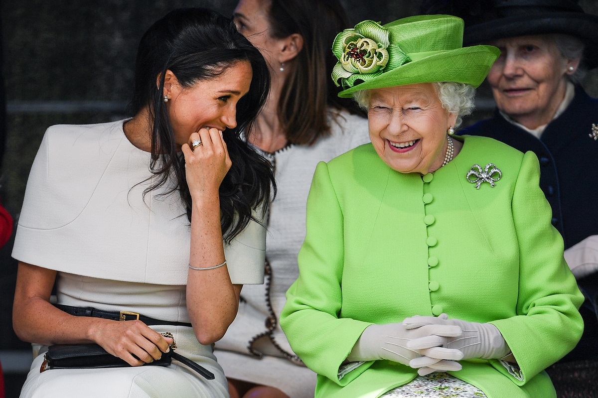 Regina Elisabeta purtând una din broșele sale celebre în formă de fundă și îmbrăcată într-un costum verde, alături de ducesa Kate îmbrăcată în alb care îi zâmbește