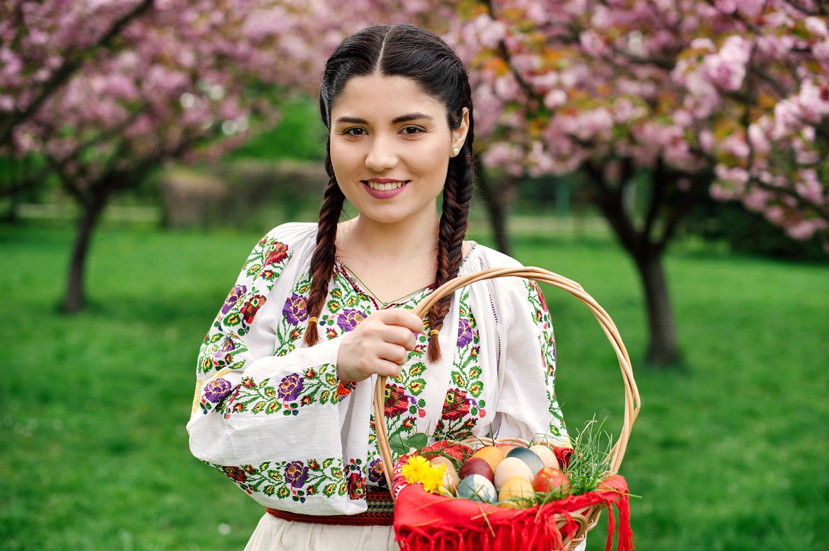 Femeie româncă într-o livadă cu cireși care ține în mână un coșuleț cu ouă vopsite pentru a prezenta cum se sărbătorește Paștele în România față de alte țări
