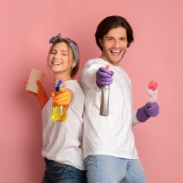 Un cuplu de tineri ce poartă mănuși de cauciuc mov și țin în mâini produse de curățenie, zâmbesc pe un fundal rozz