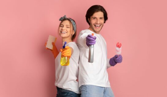 Un cuplu de tineri ce poartă mănuși de cauciuc mov și țin în mâini produse de curățenie, zâmbesc pe un fundal rozz