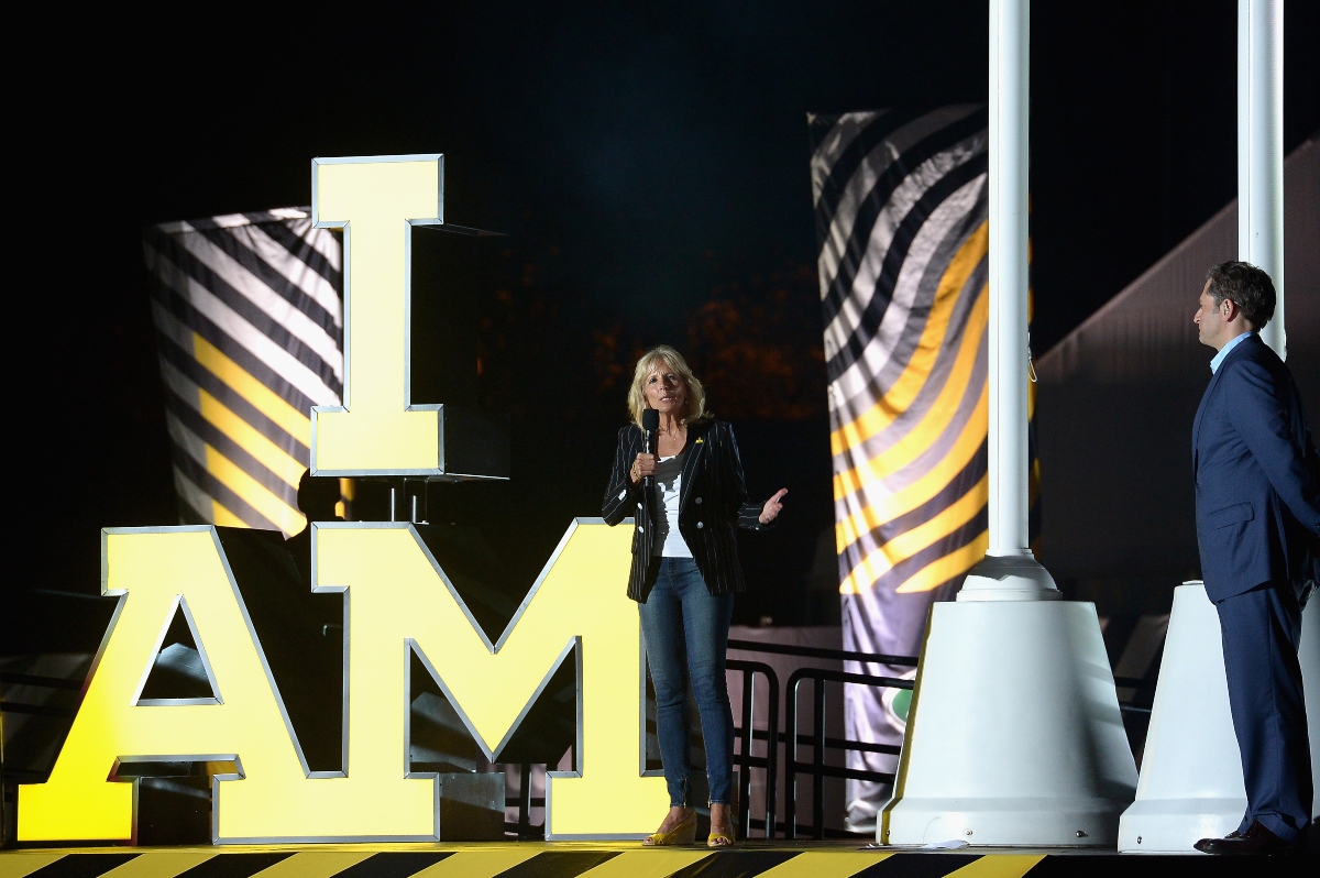 Jill Biden e pe scenă. Are în mână un microfon negru pentru că ține un discurs. Are un sacou negru cu dungi albe verticale, o pereche de blugi simpli, de culoare albastră, un top alb și niște sandale galbene, pe platformă.