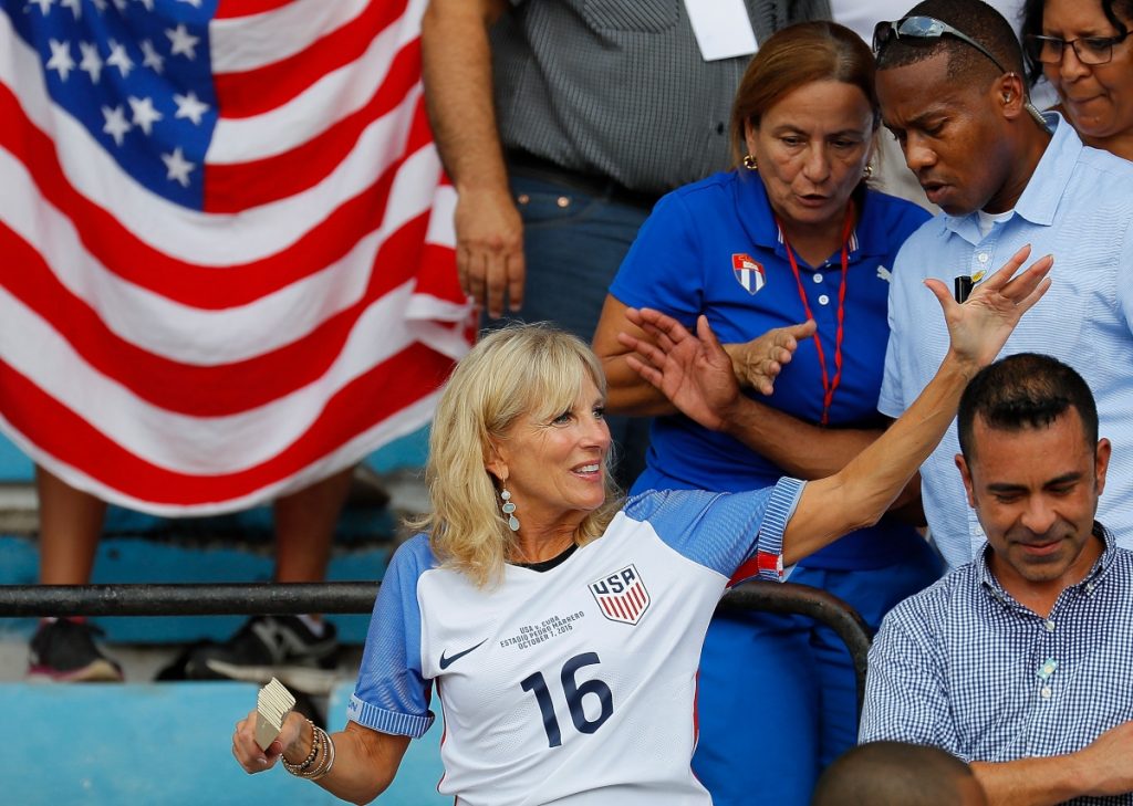 Jill Biden, în tribună la meciul de fotbal dintre SUA și Cuba din anul 2016. Poartă un tricou al echipei de fotbal, niște cercei lungi albaștri și un set de brățări la mâna dreaptă.