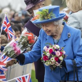 regina elisabeta într-un costum albastru cu pălărie primid flori de la copii și purtând în piept brășa sa cu safir