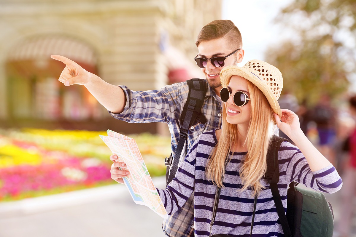 O tânără cu pălărie și ochelari de soare alături de prietenul său care îi arată una dintre cele cinci destinații sigure pentru vacanță, în timp ce tânăra ține în mână harta pe care o verifică