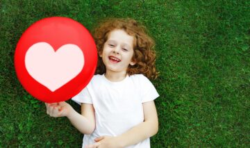 Fetiță în tricoul alb care stă ăntinsă pe iarbă în timp ce face una din acele activități creative de 1 iunie și ține în mână un balon roșu cu inimioară