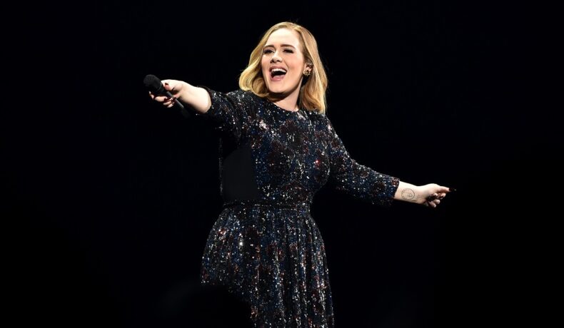 Adele pe scenă îmbrăcată într-o rochie albastră cu paiete în timp ce zâmbește și îndreaptă microfonul către public