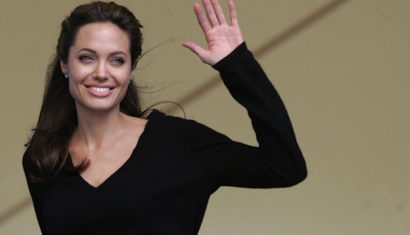 Angelina Jolie îmbrăcată într-o bluză neagră, zâmbind în timp ce face cu mâna stângă publicului în timpul conferinței live pe care a susținut-o cu muzicianul Peter Gabriel despre Africa de Sud în data e 2 iulie 2005