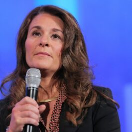 Melinda Gates, ține un microfon în mână și îmbrăcată într-o bluză cu imprimeu polka dor, peste care are un sacou negru