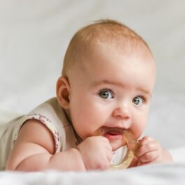 Un bebeluș cu ochișorii albaștri care ține în mână un inel de cauciuc pe care îl roade, îmbrăcată într-un body alb în timp ce stă pe așternuturi albe așteptând să primească unul dintre cele mai populare nume-de-copii-în-anul-2021