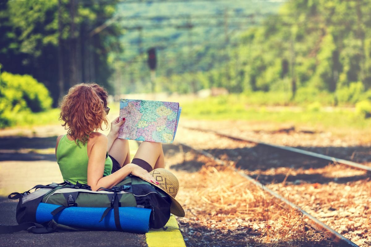 O fată, așezată pe rucsac, așteaptă trenul, într-o gară