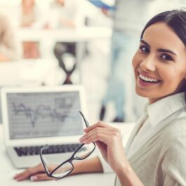O femeie tânără, lucrează la birou, în fața calculatorului, se întoarce zâmbind, cu ochearii în mână